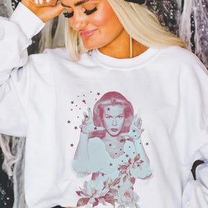 May include: White sweatshirt featuring a retro-style image of a woman with a floral and star design. The image is in shades of pink and blue. The sweatshirt has long sleeves and a crew neck.
