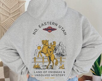 Puede incluir: Sudadera con capucha gris claro con un gráfico en la espalda. El gráfico presenta un OVNI, un vaquero a caballo y el texto "NO. EASTERN UTAH LAND OF ENIGMAS & UNSOLVED MYSTERY EST. 1886". El diseño es en tonos dorados, marrones y negros.