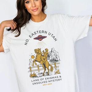 May include: White t-shirt with a graphic design featuring a cowboy riding a horse with a UFO above them. The text reads "No. Eastern Utah Land of Enigmas & Unsolved Mystery Est. 1896".