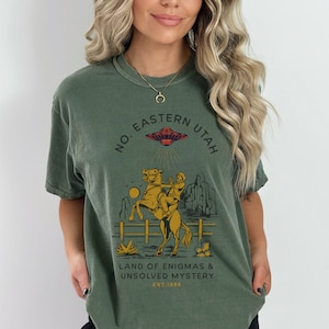 May include: Green t-shirt with a graphic print of a cowboy riding a horse with a UFO in the background. The text on the shirt reads "No. Eastern Utah Land of Enigmas & Unsolved Mystery Est. 1896"