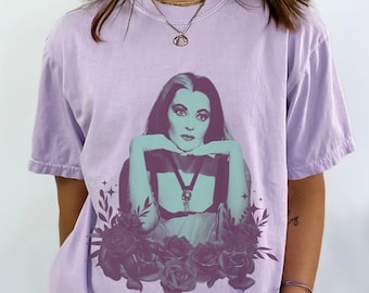 Puede incluir: Una camiseta de color lavanda con un estampado gráfico de una mujer con cabello oscuro y un vestido negro. La mujer está rodeada de rosas negras y estrellas.