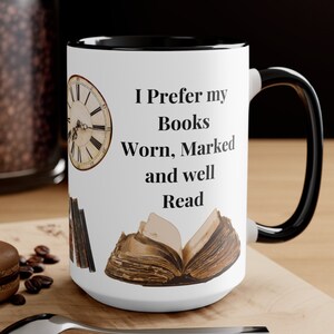 May include: White ceramic mug with a black handle and rim, featuring the text "I Prefer my Books Worn, Marked and well Read." The design includes illustrations of a clock, books, and an open book.