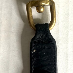 May include: A brass-toned swivel clasp with a black leather strap and a metal key ring. The clasp has a lever-operated design and a small, round stud. The key ring is attached to the leather strap with a brass-toned rivet.
