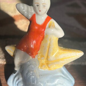 May include: A vintage porcelain figurine of a woman in an orange swimsuit, seated on a yellow starfish atop a blue wave. The figurine has a white complexion and is painted with red lips and green accents.