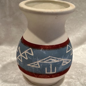 Può includere: Un piccolo vaso in ceramica con finitura bianco opaco, caratterizzato da un'ampia fascia blu al centro. La sezione blu è decorata con motivi geometrici incisi ed è delimitata da sottili strisce orizzontali rosso scuro in alto e in basso.