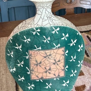 May include: A flat, teardrop-shaped ceramic vase featuring a crackle glaze finish. The neck and upper rim are cream-colored, while the body is deep green with white floral motifs and a central rectangular panel in a soft peach crackle pattern.