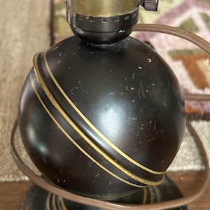 Könnte beinhalten: Eine schwarze Vintage-Tischlampe mit goldenen Akzenten. Die Lampe hat einen kugelförmigen Sockel mit zwei goldenen Bändern und einen kreisförmigen Sockel. Die Lampe hat eine Messing-Lampenfassung und ein braunes Kabel.