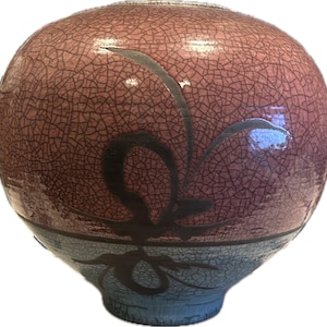 May include: A round ceramic vase featuring a distinct crackle glaze finish. The upper portion is a dusty rose color, while the lower section transitions to a light blue hue. A dark, abstract, flowing brushstroke design wraps around the center.