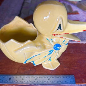 Può includere: Una fioriera a forma di anatra in ceramica vintage di colore giallo chiaro. L'anatra ha una testa grande e un becco spalancato con accenti rossi. Ha un fiocco blu e una fioriera a forma di guscio d'uovo rotto. La fioriera è lunga circa 15 cm.