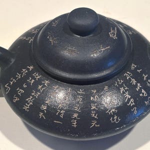 Op de afbeelding: Een donker antracietgrijze theepot met een rond lichaam, een bijpassend deksel met een centrale knop, een gebogen handvat en een korte tuit. Het buitenoppervlak van de theepot en het deksel is versierd met witte gegraveerde Chinese karakters.