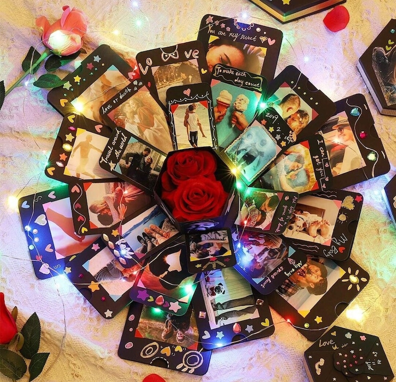 Puede incluir: Una caja explosiva hecha a mano con fotos, rosas rojas y luces de hadas. La caja negra tiene m&uacute;ltiples paneles con fotos, bordes decorativos y texto escrito a mano. El centro contiene dos rosas rojas y la caja est&aacute; rodeada de luces de cadena.
