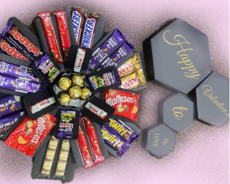 Puede incluir: Una caja de regalo hexagonal gris con texto dorado "Happy Valentines to My Love" junto a una caja explosiva abierta llena de varias barras de chocolate y caramelos, incluyendo Snickers, Lindor y Kinder.