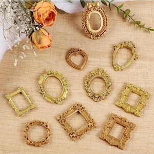May include: A collection of nine small, gold-colored decorative frames in various shapes including oval, heart, square, and rectangular. Each frame features intricate, raised relief detailing along the edges, with one oval frame topped by a bow.