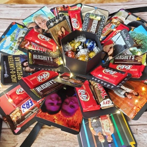 Puede incluir: Una caja explosiva negra llena de bombones y barras de chocolate, incluyendo Kit Kat, Hershey's y Twix. La caja est&aacute; rodeada de fotos y luces de hadas, creando un regalo festivo y personalizado.
