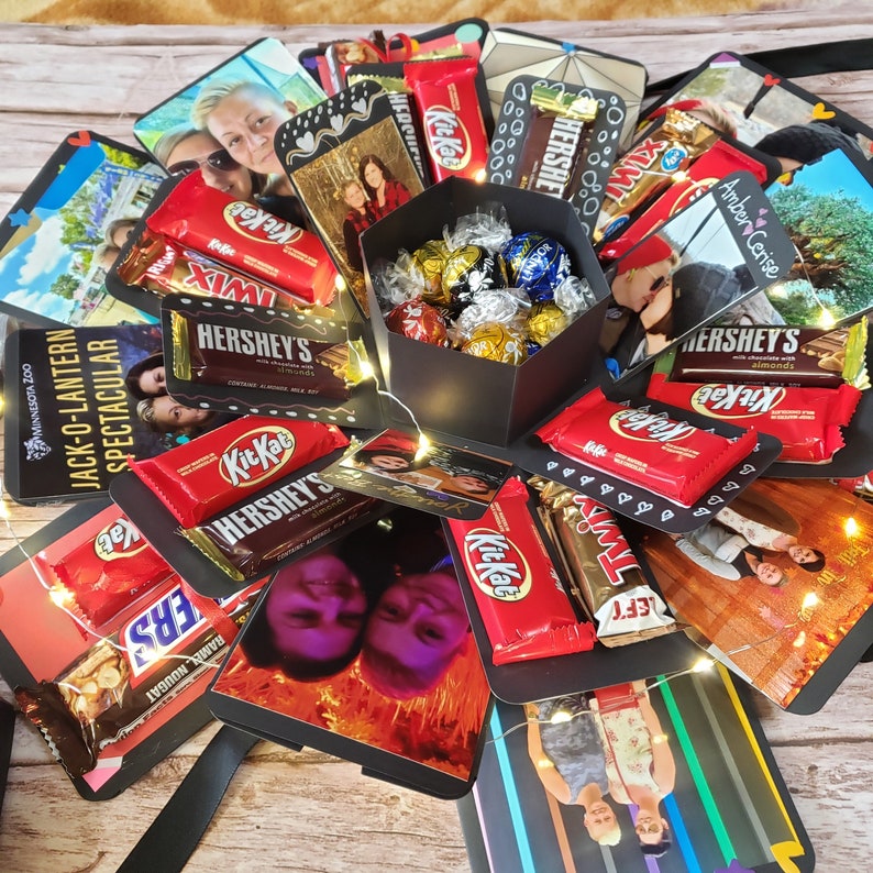 Puede incluir: Una caja explosiva negra llena de bombones y barras de chocolate, incluyendo Kit Kat, Hershey's y Twix. La caja est&aacute; rodeada de fotos y luces de hadas, creando un regalo festivo y personalizado.