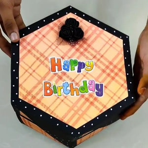 Puede incluir: Caja de regalo hexagonal con un patr&oacute;n de cuadros en tonos naranja y rojo. La caja tiene un borde negro con lunares blancos y las palabras "Happy Birthday" en letras coloridas. Un grupo de rosas negras adorna la parte superior.
