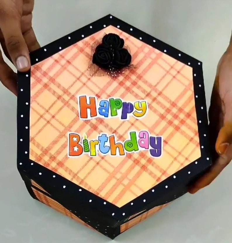 Puede incluir: Caja de regalo hexagonal con un patr&oacute;n de cuadros en tonos naranja y rojo. La caja tiene un borde negro con lunares blancos y las palabras "Happy Birthday" en letras coloridas. Un grupo de rosas negras adorna la parte superior.