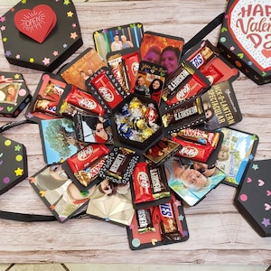 Puede incluir: Una caja explosiva negra con fotos, barras de chocolate y bombones. La caja est&aacute; decorada con estrellas y corazones de colores. En el interior hay fotos, barras Kit Kat, Hershey's y Twix. Un coraz&oacute;n rojo con "OPEN" es visible.