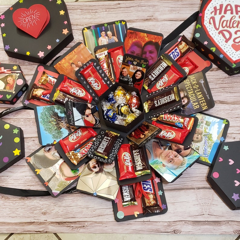Puede incluir: Una caja explosiva negra con fotos, barras de chocolate y bombones. La caja est&aacute; decorada con estrellas y corazones de colores. En el interior hay fotos, barras Kit Kat, Hershey's y Twix. Un coraz&oacute;n rojo con "OPEN" es visible.