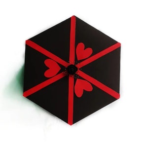 Puede incluir: Una caja de regalo hexagonal negra decorada con cintas rojas y recortes en forma de coraz&oacute;n. Una peque&ntilde;a rosa negra y puntos decorativos est&aacute;n en el centro. La caja est&aacute; sobre un fondo blanco.