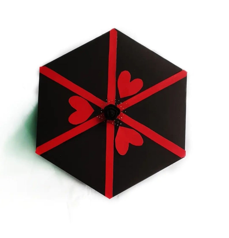 Puede incluir: Una caja de regalo hexagonal negra decorada con cintas rojas y recortes en forma de coraz&oacute;n. Una peque&ntilde;a rosa negra y puntos decorativos est&aacute;n en el centro. La caja est&aacute; sobre un fondo blanco.