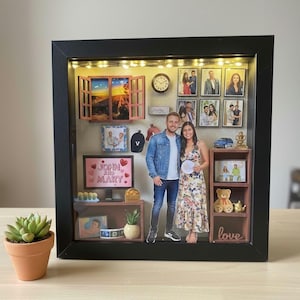 May include: A black shadow box with a variety of miniature decorative items, including photos, a clock, and a small car. The box features a couple standing in front of the display. A small potted succulent is in the foreground.