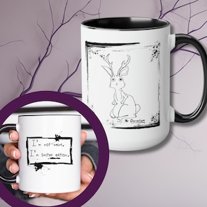 May include: White ceramic mug with a black rim and handle. The mug features a black and white illustration of a rabbit with antlers. The text "Mr. Dunlop" is written below the illustration. The mug has a black and white design with the text "I'm not weird. I'm limited edition."
