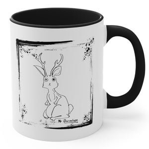 May include: White ceramic mug with a black rim and handle. The mug features a black and white illustration of a rabbit with antlers inside a distressed black frame. The text "Mr. Bunselope" is written below the illustration.