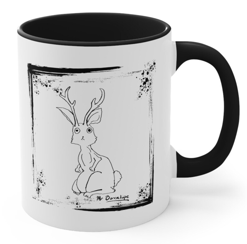 May include: White ceramic mug with a black rim and handle. The mug features a black and white illustration of a rabbit with antlers inside a distressed black frame. The text "Mr. Bunselope" is written below the illustration.