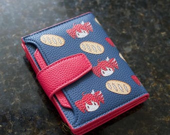 May include: A blue bi-fold wallet with a textured surface featuring a repeating pattern of small red-haired anime-style character heads and tan bread loaves. The wallet has a bright pink snap closure strap and matching pink trim along the edges.