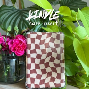 Puede incluir: Un inserto transparente para funda Kindle con un patrón de tablero de ajedrez ondulado marrón y crema. El texto "KINDLE case insert" está en la parte superior y "digital product" en la inferior. La funda se muestra frente a flores rosas y plantas verdes.