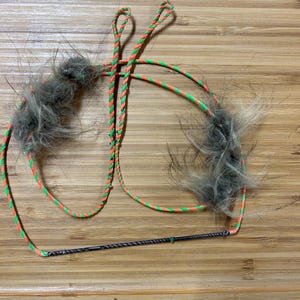 May include: A cat toy featuring two fluffy gray faux fur balls attached to a green and orange braided rope. A black, thin, flexible rod connects the two ropes. The toy is designed for interactive play.