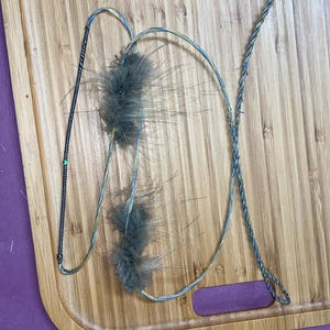 May include: A cat toy with a wooden cutting board background. The toy features a wire with two fluffy gray feather attachments and a braided rope. The cutting board is light brown with a rectangular shape and a handle cutout.