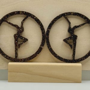 May include: Two glittery brown circular ornaments, each featuring a silhouette of a dancer in a graceful pose within the circle. The ornaments are displayed on a light-colored wooden block, set against a light wooden background. The dancer's pose is a ballet-inspired arabesque.