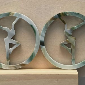 May include: Two circular pendants with a dancer silhouette inside. The pendants are made of a translucent material with green, white, and brown marbling. The dancer is in a graceful pose, arms outstretched. The pendants are displayed on a wooden surface.