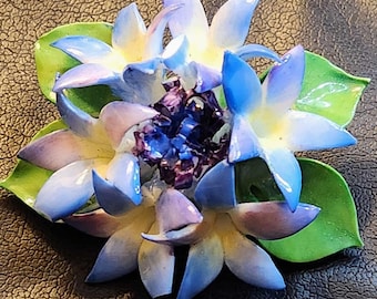May include: A blue and white porcelain flower brooch with green leaves. The flower has a purple center and is made in a realistic style.