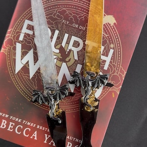 Puede incluir: Dos dagas decorativas con mangos negros en forma de dragón y detalles dorados, colocadas sobre un libro titulado "Four Winds". Las dagas tienen hojas transparentes y doradas. La portada del libro es roja con texto blanco.