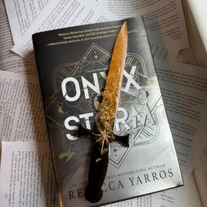 May include: A book titled "Onyx Storm" by Rebecca Yarros with a decorative dagger on top. The dagger has a black handle and a gold-colored blade with gold flecks. The book is surrounded by open pages.