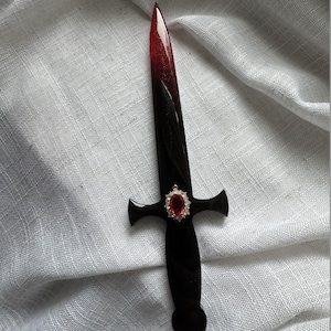 May include: A decorative dagger with a black handle and blade that fades from red to black. A red gem is set in the center of the crossguard, surrounded by a silver-colored setting. The dagger is displayed on a white, textured surface.