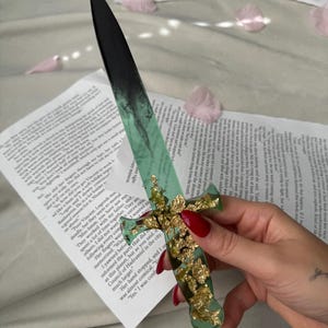 May include: A decorative dagger with a black blade and a green handle adorned with gold leaf accents. The dagger rests on a page of printed text, with pink flower petals scattered around.