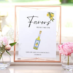 May include: A framed sign with the word "Favors" in elegant script, featuring a watercolor illustration of a limoncello bottle and lemons. The sign reads "Please Take One" and "When life gives you limoncello, celebrate."