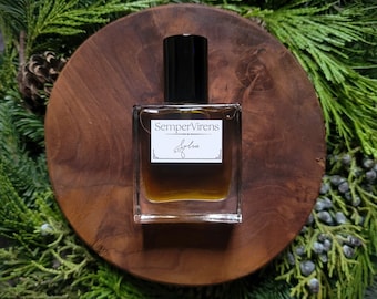 May include: A clear glass bottle with a brown liquid inside. The bottle has a black cap and a white label that says "Semper Virens" and "Sylvie". The bottle is sitting on a wooden surface.