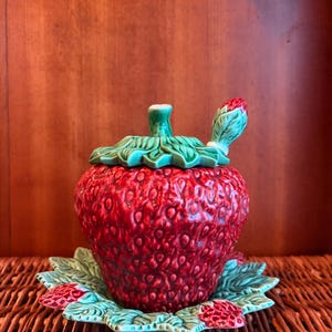 May include: A ceramic strawberry-shaped lidded jar with a matching saucer and a small spoon. The jar features a textured red surface with green leaves on the lid and base. The saucer is decorated with green leaves and small red strawberry accents.
