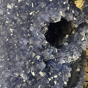 May include: A close-up of a blue crystal geode with a large opening in the center. The geode is covered in small, sparkling crystals.