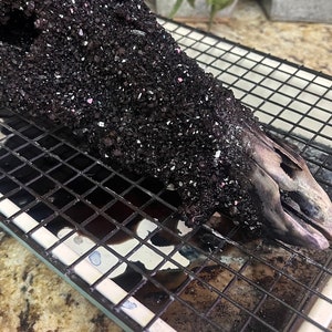 May include: A black metal cooling rack with a black, crystal-like substance on top. The substance is in a large, irregular shape and appears to be melting. The rack is sitting on a white baking sheet.