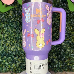 May include: A purple Stanley Quencher H2.0 tumbler with a handle, featuring a design of Easter bunnies, bows, and stars. The tumbler has a capacity of 40 oz (1.18 L) and includes the text "All Day Hydration".