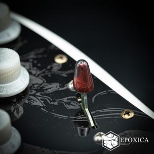 Puede incluir: Un selector de pastillas de guitarra de color rojo oscuro brillante montado sobre un golpeador negro con un patrón floral blanco. El golpeador presenta un borde blanco, tornillos metálicos y el logotipo blanco EPOXICA Guitars.