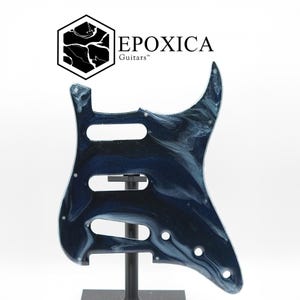 Puede incluir: Un golpeador de guitarra azul y blanco veteado con tres orificios de control y una ranura para el brazo del trémolo. El golpeador está montado sobre un soporte negro. El logotipo "EPOXICA Guitars" está en la esquina superior izquierda.