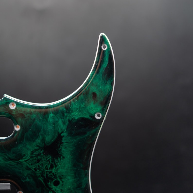 May include: A guitar pickguard with a swirling green and black pattern. The pickguard has a white edge and several pre-drilled holes for mounting. The design resembles a marbled or stone-like texture, adding a unique aesthetic to the instrument.