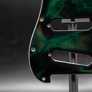 May include: Close-up of a guitar pickguard with a swirling green and black pattern. The pickguard has three cutouts for pickups and several screw holes. The edges are white, and the pickguard is mounted on a stand.